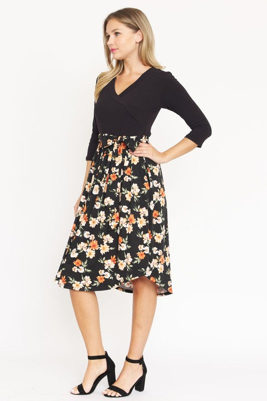 A woman in a Surplice Floral Sash Midi Dress, comprised of 95% Polyester and 5% Spandex, stands against a plain white backdrop. The knee-length dress, designed in navy blue with a floral skirt and wrapped bodice, showcases elegant craftsmanship from the United States.