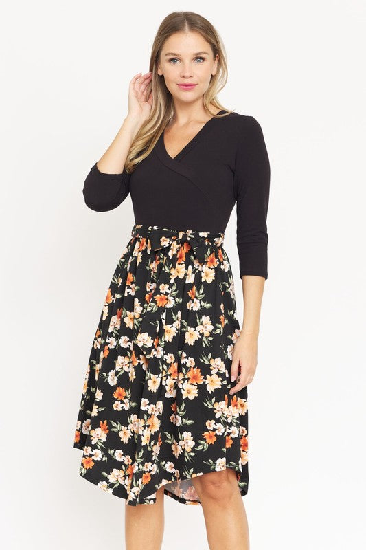 A woman in a Surplice Floral Sash Midi Dress, comprised of 95% Polyester and 5% Spandex, stands against a plain white backdrop. The knee-length dress, designed in navy blue with a floral skirt and wrapped bodice, showcases elegant craftsmanship from the United States.