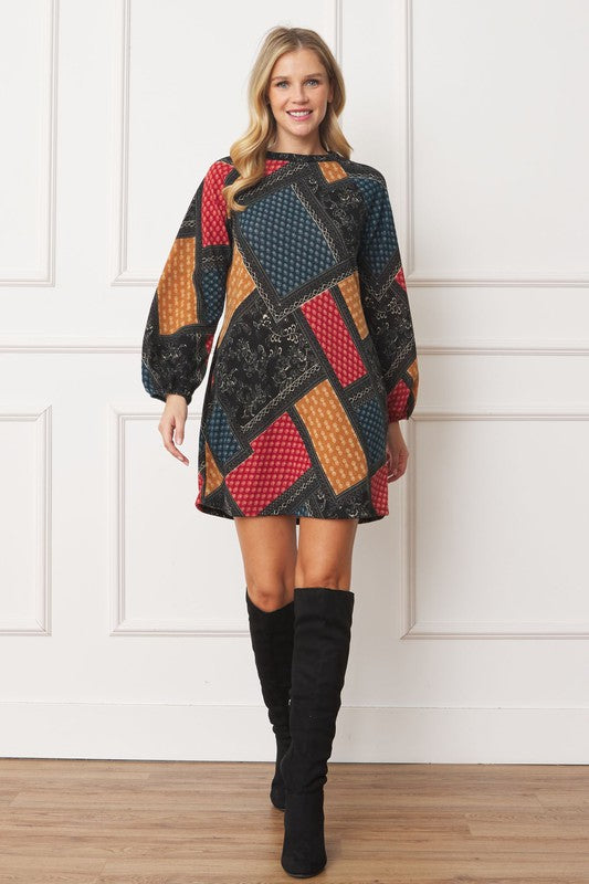A woman wears the Bishop Sleeve Multi Patch Pattern Mini Dress, featuring chic long bishop sleeves and a multicolored patchwork design with panels of red, blue, black, and yellow. She stands against a plain white background.