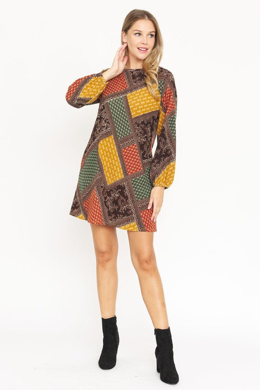 A woman wears the Bishop Sleeve Multi Patch Pattern Mini Dress, featuring chic long bishop sleeves and a multicolored patchwork design with panels of red, blue, black, and yellow. She stands against a plain white background.