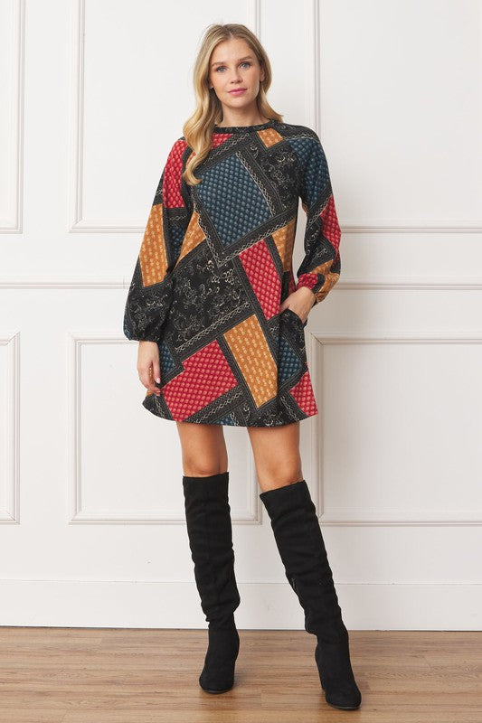A woman wears the Bishop Sleeve Multi Patch Pattern Mini Dress, featuring chic long bishop sleeves and a multicolored patchwork design with panels of red, blue, black, and yellow. She stands against a plain white background.