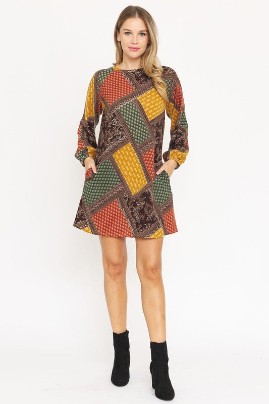 A woman wears the Bishop Sleeve Multi Patch Pattern Mini Dress, featuring chic long bishop sleeves and a multicolored patchwork design with panels of red, blue, black, and yellow. She stands against a plain white background.