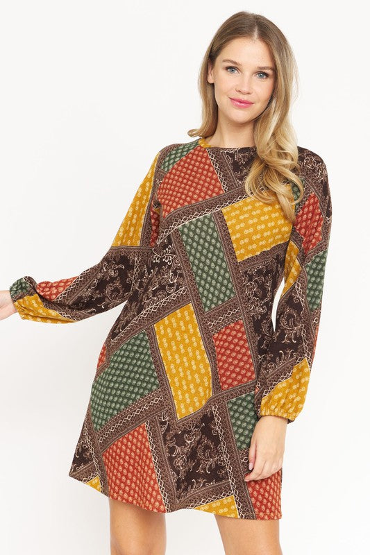 A woman wears the Bishop Sleeve Multi Patch Pattern Mini Dress, featuring chic long bishop sleeves and a multicolored patchwork design with panels of red, blue, black, and yellow. She stands against a plain white background.