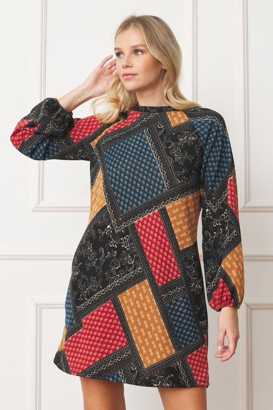 A woman wears the Bishop Sleeve Multi Patch Pattern Mini Dress, featuring chic long bishop sleeves and a multicolored patchwork design with panels of red, blue, black, and yellow. She stands against a plain white background.