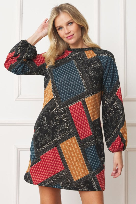 A woman wears the Bishop Sleeve Multi Patch Pattern Mini Dress, featuring chic long bishop sleeves and a multicolored patchwork design with panels of red, blue, black, and yellow. She stands against a plain white background.