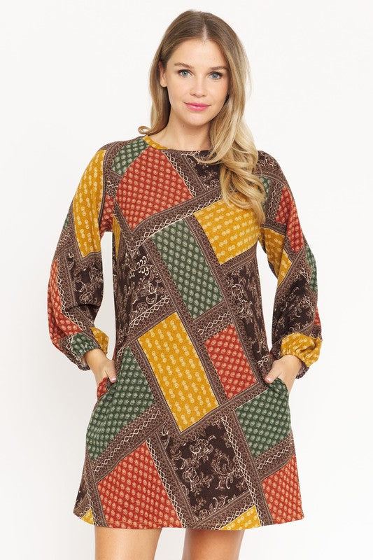 A woman wears the Bishop Sleeve Multi Patch Pattern Mini Dress, featuring chic long bishop sleeves and a multicolored patchwork design with panels of red, blue, black, and yellow. She stands against a plain white background.