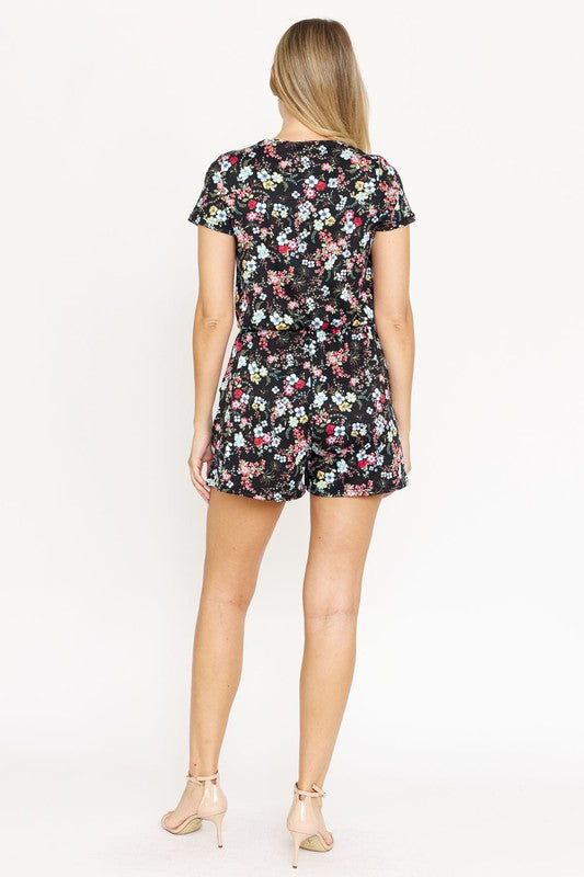 A woman is smiling at the camera, wearing a stylish Surplice Drawstring Floral Romper with short sleeves and a V-neckline.