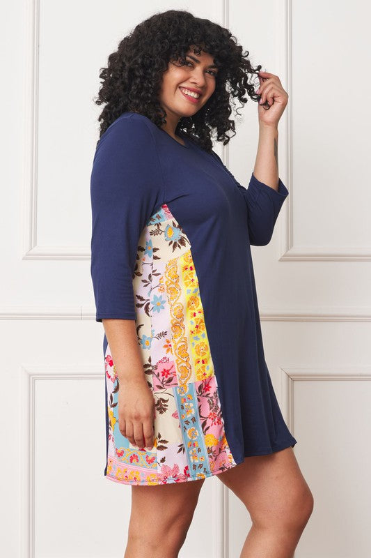 A woman with curly hair wearing a Plus Side Contrast A Line Tunic Dress in navy blue, featuring side contrast floral panels, stands in front of a white paneled wall.