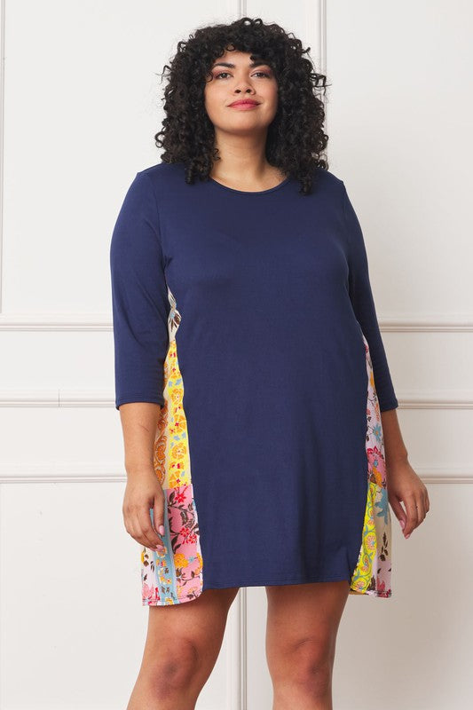 A woman with curly hair wearing a Plus Side Contrast A Line Tunic Dress in navy blue, featuring side contrast floral panels, stands in front of a white paneled wall.