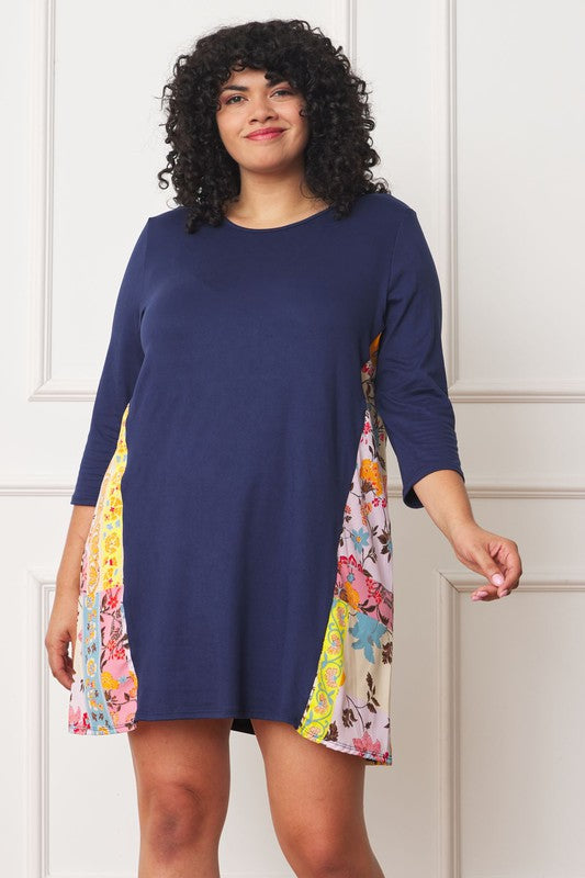 A woman with curly hair wearing a Plus Side Contrast A Line Tunic Dress in navy blue, featuring side contrast floral panels, stands in front of a white paneled wall.