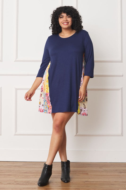 A woman with curly hair wearing a Plus Side Contrast A Line Tunic Dress in navy blue, featuring side contrast floral panels, stands in front of a white paneled wall.