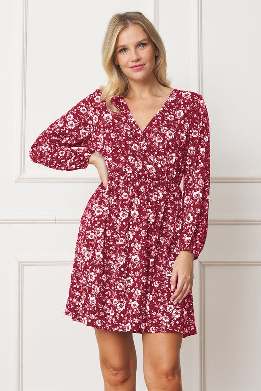 A woman stands against a white paneled wall, wearing the V-Neck Flowy Floral Mini Dress in a maroon floral pattern. The dress features long sleeves and falls gracefully above the knee. She is smiling with one hand on her hip. Made from 95% polyester and 5% spandex, this elegant dress is proudly crafted in the United States.