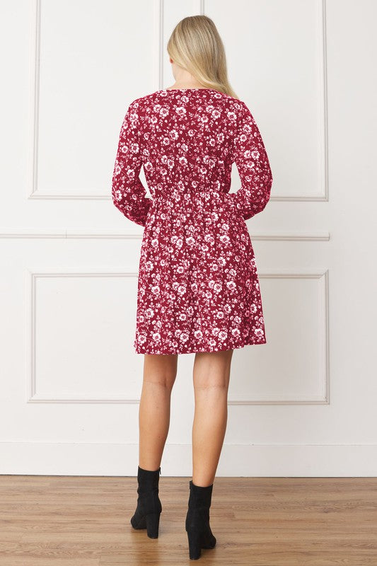 A woman stands against a white paneled wall, wearing the V-Neck Flowy Floral Mini Dress in a maroon floral pattern. The dress features long sleeves and falls gracefully above the knee. She is smiling with one hand on her hip. Made from 95% polyester and 5% spandex, this elegant dress is proudly crafted in the United States.