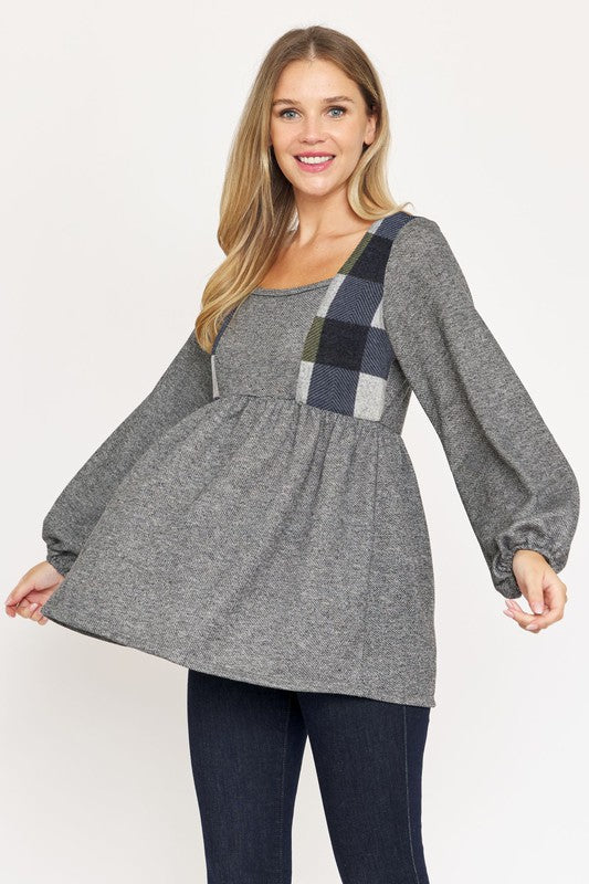 A person with long hair is standing, wearing a grey long-sleeved Terry Plaid Side Contrast Baby Doll Tunic featuring black and red plaid details, along with dark pants against a plain white background.