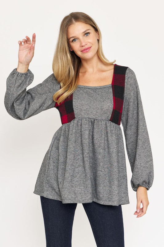 A woman stands against a plain background, smiling and touching the side of her head. She is wearing a gray Terry Plaid Side Contrast Baby Doll Tunic with checkered strap accents and dark pants, proudly made in the United States.