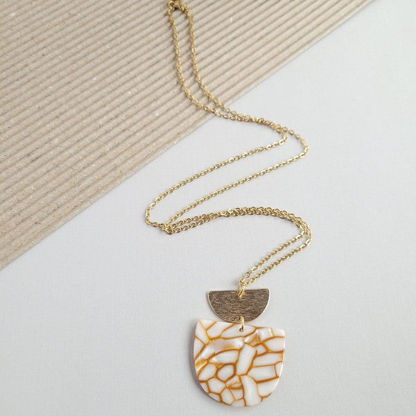 The Harper Pumpkin Spice Necklace, a gold-plated chain adorned with a pendant showcasing cracked white and orange acrylic hues along with a small gold half-moon detail, is elegantly placed on a textured, two-tone background.