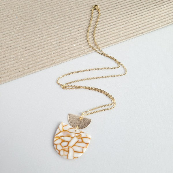 The Harper Pumpkin Spice Necklace, a gold-plated chain adorned with a pendant showcasing cracked white and orange acrylic hues along with a small gold half-moon detail, is elegantly placed on a textured, two-tone background.