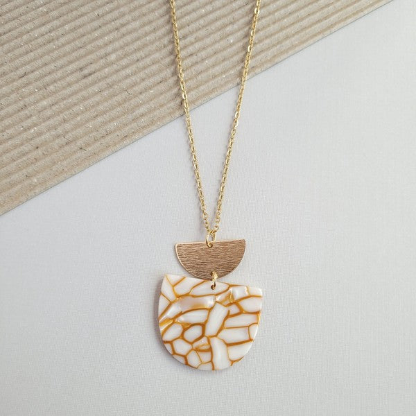 The Harper Pumpkin Spice Necklace, a gold-plated chain adorned with a pendant showcasing cracked white and orange acrylic hues along with a small gold half-moon detail, is elegantly placed on a textured, two-tone background.