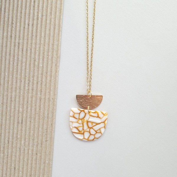 The Harper Pumpkin Spice Necklace, a gold-plated chain adorned with a pendant showcasing cracked white and orange acrylic hues along with a small gold half-moon detail, is elegantly placed on a textured, two-tone background.