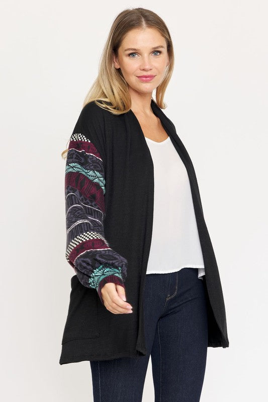 A woman stands against a white background, dressed in a black Bishop Sleeve Open Cardigan With Pockets that has colorful patterned sleeves. She pairs it with a white top and dark jeans. This stylish piece is made in the United States.