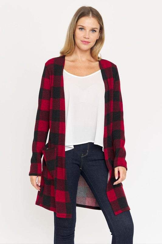 A woman in a Long Buffalo Plaid Open Cardigan, featuring a striking red design and crafted from soft polyester spandex, stands against a plain background. She pairs it with a white top and dark jeans, facing the camera with her hands in front pockets. Made in the United States, this look combines comfort and style effortlessly.