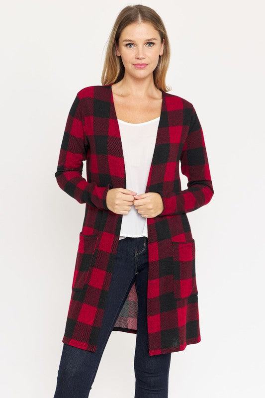 A woman in a Long Buffalo Plaid Open Cardigan, featuring a striking red design and crafted from soft polyester spandex, stands against a plain background. She pairs it with a white top and dark jeans, facing the camera with her hands in front pockets. Made in the United States, this look combines comfort and style effortlessly.