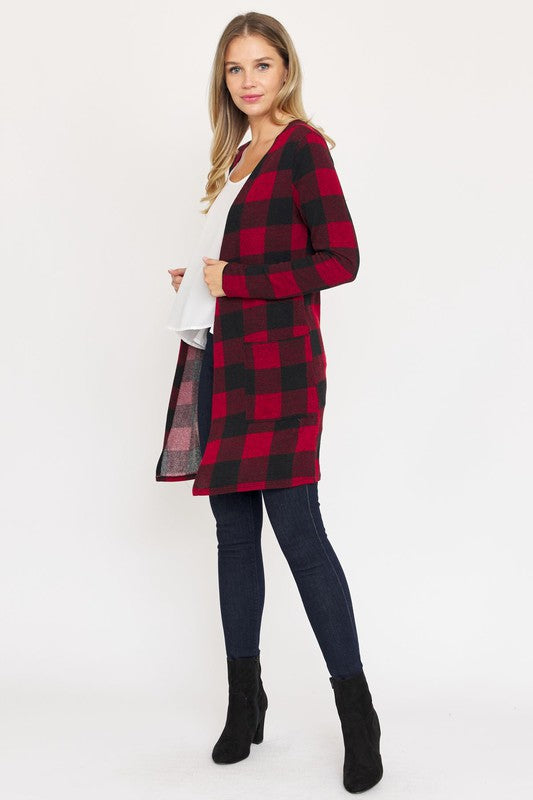 A woman in a Long Buffalo Plaid Open Cardigan, featuring a striking red design and crafted from soft polyester spandex, stands against a plain background. She pairs it with a white top and dark jeans, facing the camera with her hands in front pockets. Made in the United States, this look combines comfort and style effortlessly.