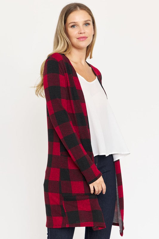 A woman in a Long Buffalo Plaid Open Cardigan, featuring a striking red design and crafted from soft polyester spandex, stands against a plain background. She pairs it with a white top and dark jeans, facing the camera with her hands in front pockets. Made in the United States, this look combines comfort and style effortlessly.