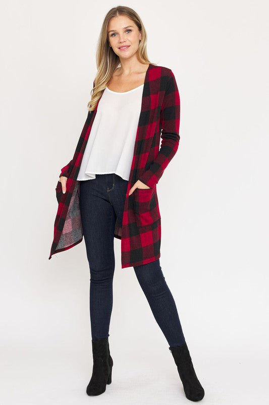 A woman in a Long Buffalo Plaid Open Cardigan, featuring a striking red design and crafted from soft polyester spandex, stands against a plain background. She pairs it with a white top and dark jeans, facing the camera with her hands in front pockets. Made in the United States, this look combines comfort and style effortlessly.