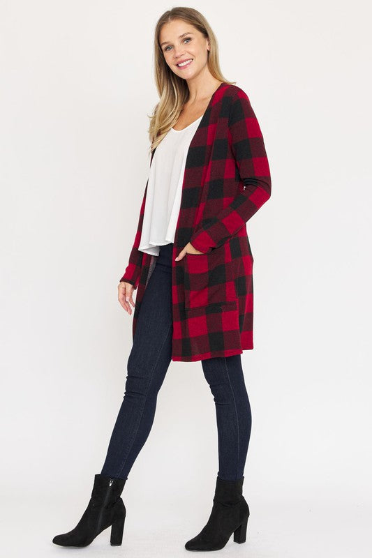 A woman in a Long Buffalo Plaid Open Cardigan, featuring a striking red design and crafted from soft polyester spandex, stands against a plain background. She pairs it with a white top and dark jeans, facing the camera with her hands in front pockets. Made in the United States, this look combines comfort and style effortlessly.