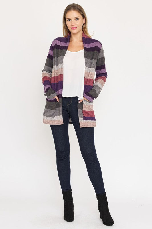 A person stands wearing the Stripe Elbow Patch Cardigan, a stylish garment featuring various shades of purple, gray, and pink stripes over a white top. With hands tucked into the cardigan's pockets, they showcase its chic design made from Polyester Spandex. The background is plain white.