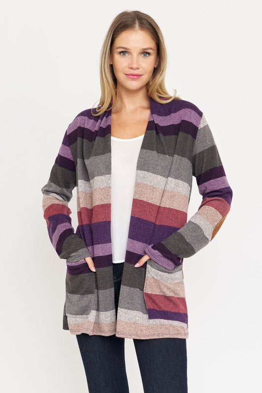 A person stands wearing the Stripe Elbow Patch Cardigan, a stylish garment featuring various shades of purple, gray, and pink stripes over a white top. With hands tucked into the cardigan's pockets, they showcase its chic design made from Polyester Spandex. The background is plain white.