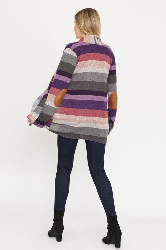 A person stands wearing the Stripe Elbow Patch Cardigan, a stylish garment featuring various shades of purple, gray, and pink stripes over a white top. With hands tucked into the cardigan's pockets, they showcase its chic design made from Polyester Spandex. The background is plain white.