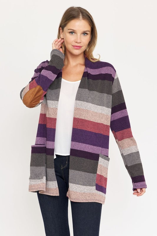 A person stands wearing the Stripe Elbow Patch Cardigan, a stylish garment featuring various shades of purple, gray, and pink stripes over a white top. With hands tucked into the cardigan's pockets, they showcase its chic design made from Polyester Spandex. The background is plain white.