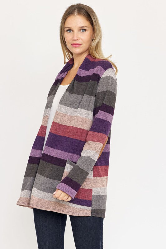 A person stands wearing the Stripe Elbow Patch Cardigan, a stylish garment featuring various shades of purple, gray, and pink stripes over a white top. With hands tucked into the cardigan's pockets, they showcase its chic design made from Polyester Spandex. The background is plain white.