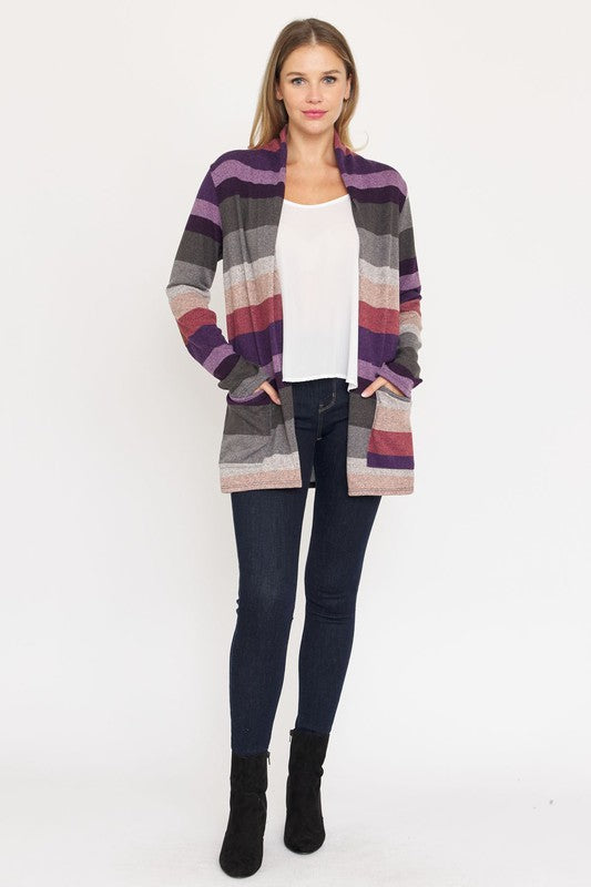 A person stands wearing the Stripe Elbow Patch Cardigan, a stylish garment featuring various shades of purple, gray, and pink stripes over a white top. With hands tucked into the cardigan's pockets, they showcase its chic design made from Polyester Spandex. The background is plain white.