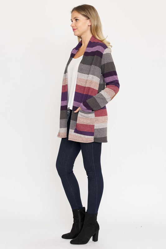 A person stands wearing the Stripe Elbow Patch Cardigan, a stylish garment featuring various shades of purple, gray, and pink stripes over a white top. With hands tucked into the cardigan's pockets, they showcase its chic design made from Polyester Spandex. The background is plain white.