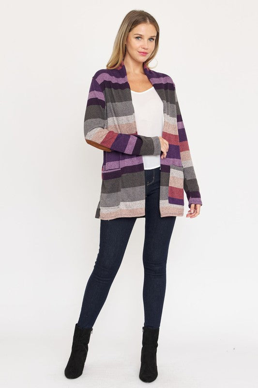 A person stands wearing the Stripe Elbow Patch Cardigan, a stylish garment featuring various shades of purple, gray, and pink stripes over a white top. With hands tucked into the cardigan's pockets, they showcase its chic design made from Polyester Spandex. The background is plain white.