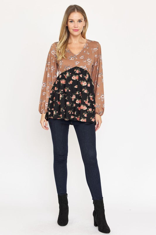 A woman stands, wearing a V Neck Tiered Baby Doll Top with a brown and black floral pattern made from polyester spandex fabric. She pairs it with dark blue skinny jeans and black ankle boots. With her long blonde hair flowing and a smile on her face, she embodies effortless style. This top is proudly made in the USA.