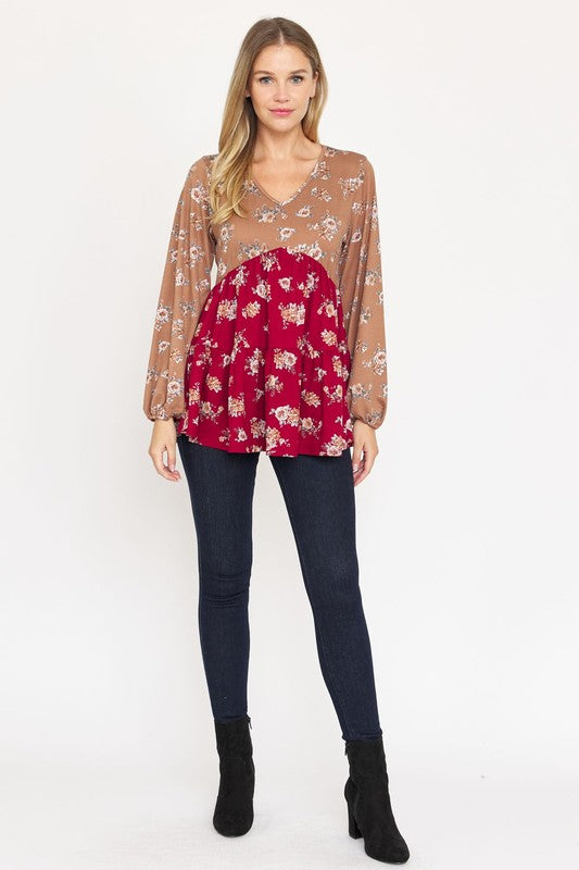 A woman stands, wearing a V Neck Tiered Baby Doll Top with a brown and black floral pattern made from polyester spandex fabric. She pairs it with dark blue skinny jeans and black ankle boots. With her long blonde hair flowing and a smile on her face, she embodies effortless style. This top is proudly made in the USA.