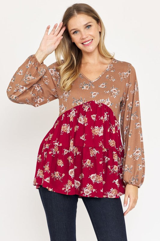 A smiling woman with long hair waves while wearing a V Neck Tiered Baby Doll Top with tan and red sections, paired with dark pants. Made in USA.