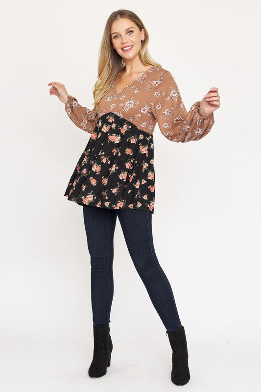 A woman stands, wearing a V Neck Tiered Baby Doll Top with a brown and black floral pattern made from polyester spandex fabric. She pairs it with dark blue skinny jeans and black ankle boots. With her long blonde hair flowing and a smile on her face, she embodies effortless style. This top is proudly made in the USA.