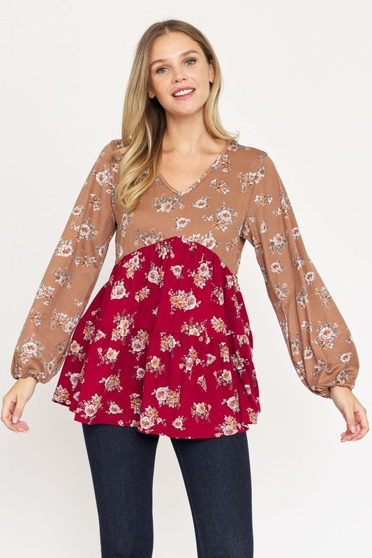 A woman stands, wearing a V Neck Tiered Baby Doll Top with a brown and black floral pattern made from polyester spandex fabric. She pairs it with dark blue skinny jeans and black ankle boots. With her long blonde hair flowing and a smile on her face, she embodies effortless style. This top is proudly made in the USA.