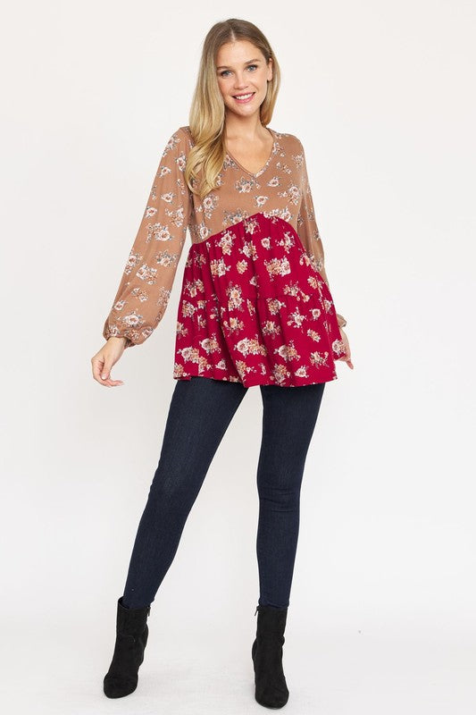 A woman stands, wearing a V Neck Tiered Baby Doll Top with a brown and black floral pattern made from polyester spandex fabric. She pairs it with dark blue skinny jeans and black ankle boots. With her long blonde hair flowing and a smile on her face, she embodies effortless style. This top is proudly made in the USA.