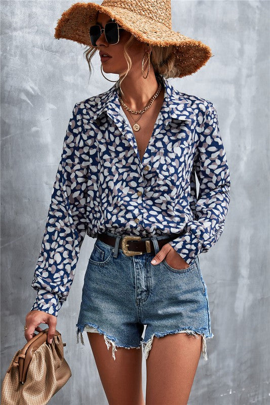 A person wearing a wide-brimmed straw hat, sunglasses, a patterned Women's Long Sleeve Blouse made from polyester spandex fabric, denim shorts, and holding a brown clutch bag stands against a gray background.