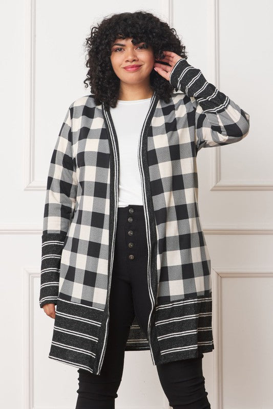 A person with curly hair is wearing a Long Buffalo Plaid Stripe Trim Open Cardigan in black and white over a white shirt, paired with black high-waisted pants with buttons. They stand in front of a white paneled wall.