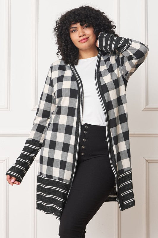 A person with curly hair is wearing a Long Buffalo Plaid Stripe Trim Open Cardigan in black and white over a white shirt, paired with black high-waisted pants with buttons. They stand in front of a white paneled wall.