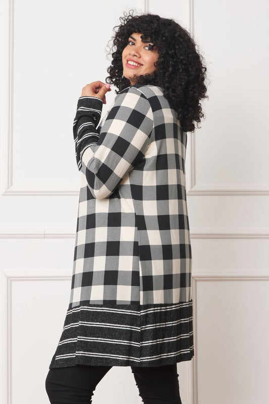 A person with curly hair is wearing a Long Buffalo Plaid Stripe Trim Open Cardigan in black and white over a white shirt, paired with black high-waisted pants with buttons. They stand in front of a white paneled wall.