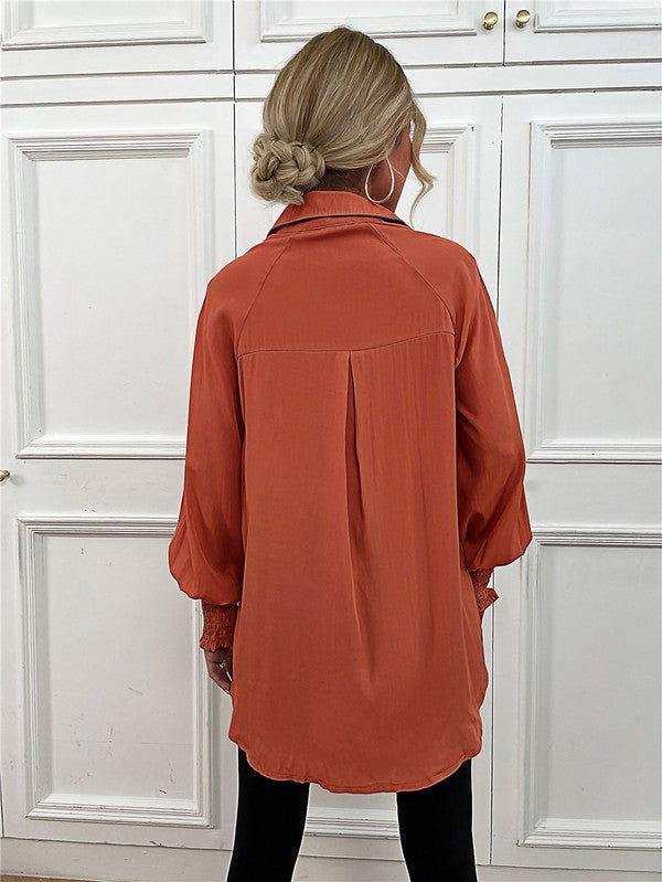 A woman wearing a Women's Long Sleeve Button Down Bluse with long puffy sleeves, made from 100% polyester, and layered necklaces stands in front of a white paneled wall.