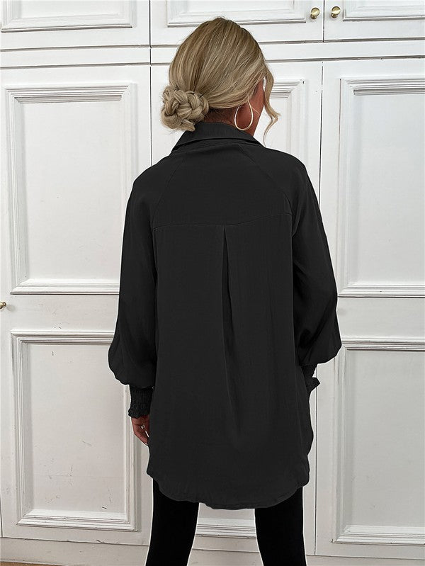 A woman wearing a Women's Long Sleeve Button Down Bluse with long puffy sleeves, made from 100% polyester, and layered necklaces stands in front of a white paneled wall.